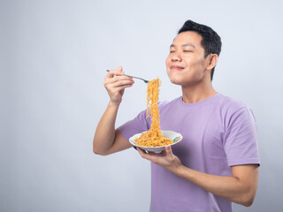 Young Asian man in a lavender t-shirt enthusiastically eating instant noodles from a bowl with a...