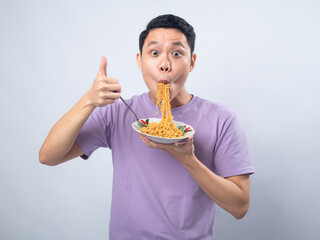 Young Asian man in a lavender t-shirt enthusiastically eating instant noodles from a bowl with a...