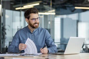 A smiling young man sits in the office at a table in front of a laptop, makes notes in documents, works with data