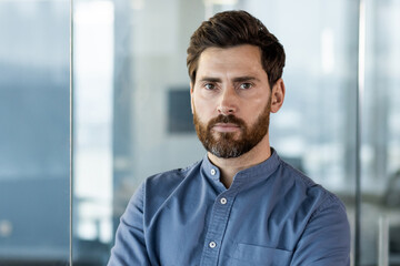 Obraz premium Close-up portrait of a young serious businessman in a blue shirt standing inside the office and looking confidently at the camera