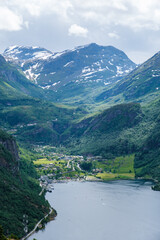 A stunning aerial view of a Norwegian fjord valley, showcasing snow-capped mountains, lush green vegetation, and a winding river leading into a picturesque village. Geiranger fjord Norway