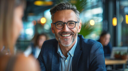 Fototapeta premium A cheerful middle-aged man with glasses smiles while sitting in a modern cafe, creating a welcoming atmosphere.