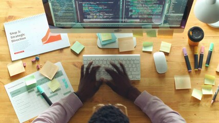 Black male programmer typing code on computer at office desk with business papers and sticky notes during workday in IT company. Time lapse, top down shot