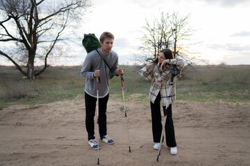 couple, exhausted after a long day of trekking, take a break on a dirt path. They are both leaning on their trekking poles, and the woman is wiping her brow