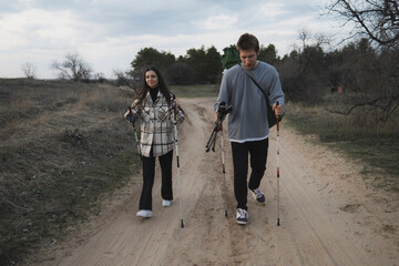 couple treks through countryside using poles. Trail surrounded by grassy fields, trees, distant cityscape in background