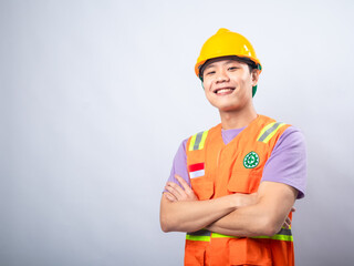 Young Asian man in a lavender t-shirt wearing an orange safety vest and yellow hard hat, standing with arms crossed and smiling. Studio shot on a plain background highlighting his work attire