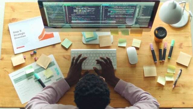 African American man typing programming code on computer at office desk with business documents and sticky notes on it as working in IT company. Directly above view