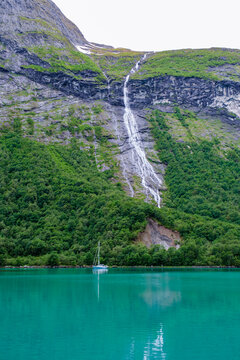 A sailboat rests serenely on the turquoise waters of a Norwegian lake, with a dramatic waterfall cascading down the steep mountainside behind it. Norangsfjorden Norangdal Urke Norway