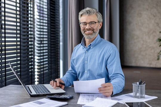 Smiling mature businessman working on a laptop while sitting at his desk in a modern office environment. Holding papers and surrounded by documents.