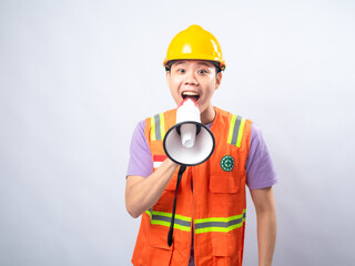 Young Asian man in an orange safety vest and yellow hard hat, holding a megaphone and shouting, isolated on a white background. The image conveys urgency, communication
