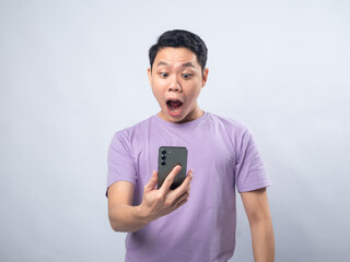 Young Asian man in a lavender t-shirt looking surprised and excited while waving at his smartphone....