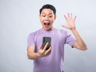 Young Asian man in a lavender t-shirt looking surprised and excited while waving at his smartphone....
