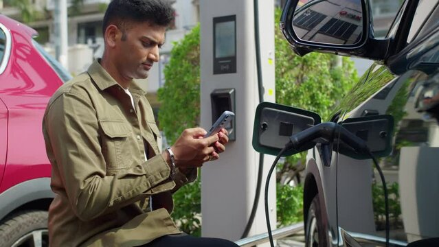 Side view of male Indian driver checking application on smartphone when inserting charging cable in port at city station