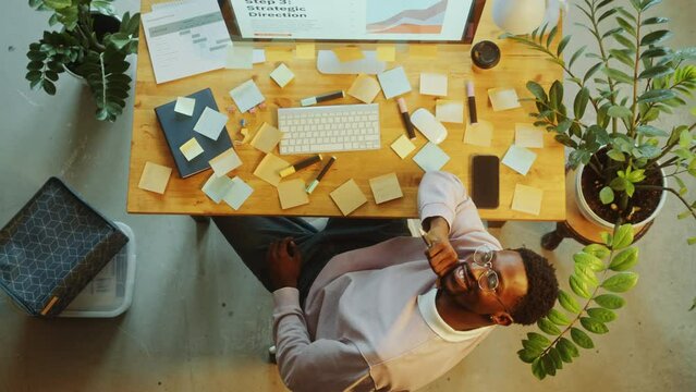 Young African American businessman in casual wear sitting at desk with computer and sticky notes on it, posing on camera and smiling during workday in office. Top down view