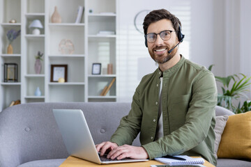 Fototapeta premium Smiling man using a laptop and wearing a headset while working from home, participating in a video call.