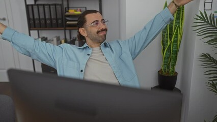 Young man with moustache in an office setting, working on a computer, stretching and expressing positive emotions surrounded by indoor plants and office supplies.