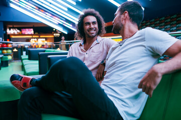 Two handsome male friends smiling and looking each other while relaxing on comfortable couch in...