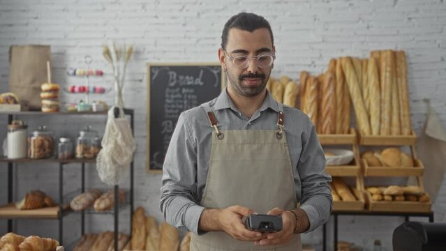 Young hispanic man in apron using payment terminal in cozy bakery shop with bread on shelves and jars of cookies and breadsticks