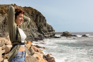 A woman stands on a rocky shoreline, smiling and looking out at the ocean