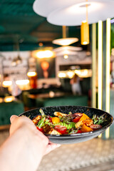 Close-up of a hand holding a colorful salad on a black plate in a restaurant setting