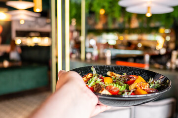 Close-up of a hand holding a colorful salad on a black plate in a restaurant setting