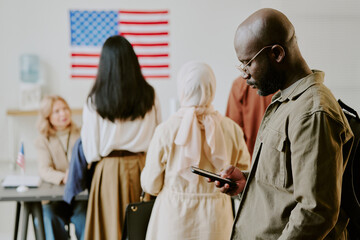 Young African American man scrolling web page on smartphone while waiting in line at polling station on election day