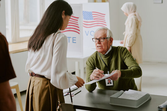 Senior Caucasian electoral district worker sitting at desk at polling station explaining young Asian woman how to fill in ballot paper