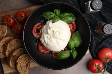 Delicious burrata cheese, tomatoes, basil and bread in bowl on brown table, flat lay