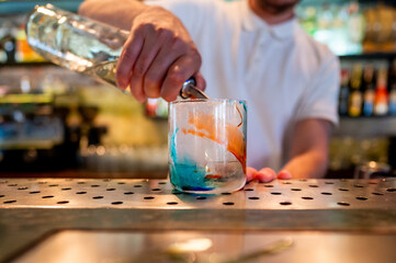 Close-up of a hand pouring a clear liquid into a glass with blue and orange streaks on a bar counter.