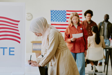 Young biracial Muslim woman wearing hijab voting for candidate in ballot paper at polling station...