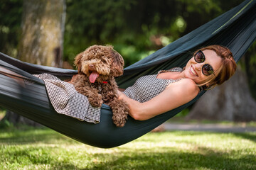 Woman relaxes in hammock with her fluffy brown Maltipoo dog on sunny day. Both looking content and...