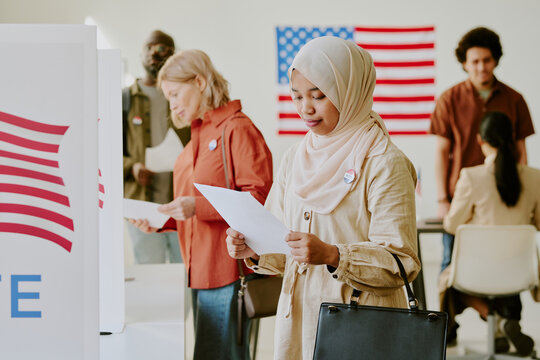 Medium shot of young Muslim woman in hijab standing at polling station looking at list of presidential candidates