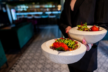 close-up of a person in a black outfit holding two white plates with gourmet dishes, featuring vibrant green avocado and red strawberries, in a dimly lit restaurant setting
