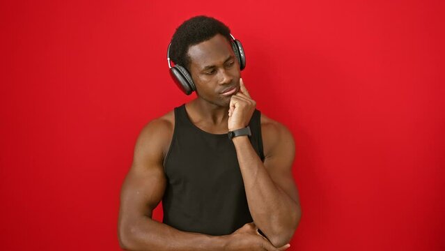 Pensive young african american man, earnestly questioning his beliefs over his chin, seriously engrossed listening to music, a troubling idea on his face, isolated against a red wall.