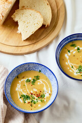Two bowls of creamy potato soup, spoons, napkin and wooden cutting board with a loaf of bread and cut pieces. The surface of the soup is decorated with cream, croutons, parsley and ground red pepper.