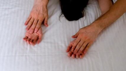 Man is stroking woman's hands lying in bed with white linen making love, hands close-up. Sex, intimate life, pleasure and love concept. Two lovers having a sex holding hands, sensual gesture.