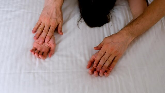 Man is stroking woman's hands lying in bed with white linen making love, hands close-up. Sex, intimate life, pleasure and love concept. Two lovers having a sex holding hands, sensual gesture.