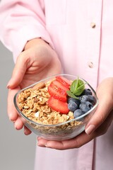 Woman holding bowl of tasty granola with berries, nuts and mint on light grey background, closeup