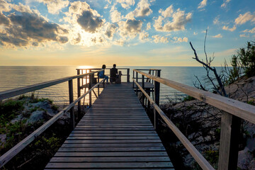 Naklejka premium A perspective of a wooden boardwalk across a dune. In the distance there are dark silhouettes of people sitting, admiring a beautiful sunset over the sea.