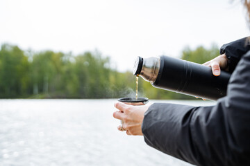 A person is pouring water from a bottle into a cup, with a sleeve on their thumb. The gesture is graceful against the backdrop of the sky, grass, and lake in the serene landscape