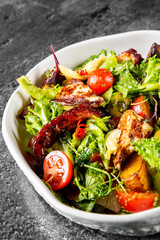 Fresh mixed salad with grilled octopus, cherry tomatoes, and greens in a white bowl on a dark textured background