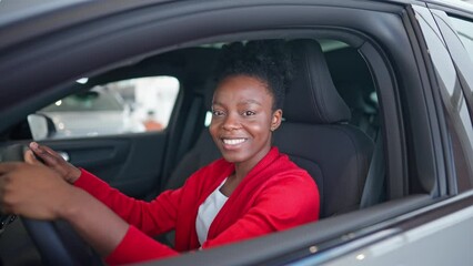 Adorable African American woman sitting in luxury car. Satisfied girl holding hands on steering wheel while imagining driving. Positively looking at camera and cheerfully smiling after test drive.