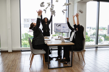 Business team in professional attire celebrating success by throwing money in modern office with large windows, laptops on desk. Emphasis on teamwork, achievement, and workplace celebration.