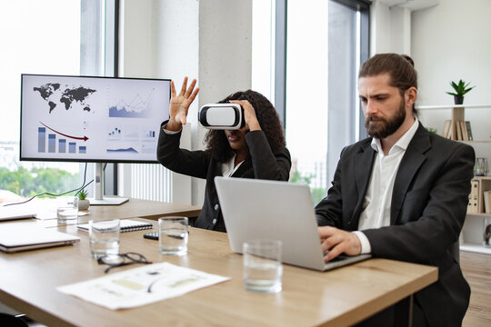 Business colleagues in formal attire at office table, one using virtual reality headset while the other works on laptop. Charts and graphs displayed on monitor in background, modern office setting.
