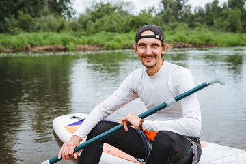 A man enjoys paddleboarding on a calm river, surrounded by nature. Outdoor water sports provide a serene and leisurely activity for an active lifestyle, making him happy