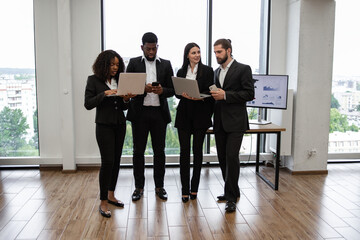 Fototapeta premium Business team of four professionals collaborating in modern office, using laptops and smartphone. Office environment features large windows with city view and presentation screen displaying charts.
