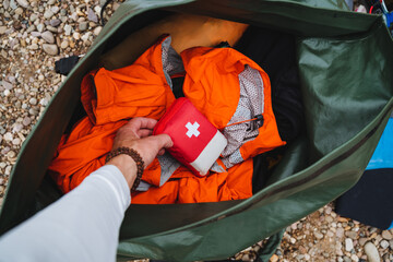 A person carrying a first aid kit in a bag filled with clothes, sports gear, hiking equipment, an orange bag, and personal protective gear, standing amidst grass and trees © Aleksey
