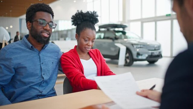 View of multi-ethnic people sitting at table and signing car purchase documentation in auto showroom. Manager or salesman explaining something to his customers. In background visible vehicle.