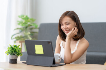 Fototapeta premium Smiling woman working on a tablet at home with a cup of coffee, surrounded by green plants, enjoying a cozy and productive work environment.