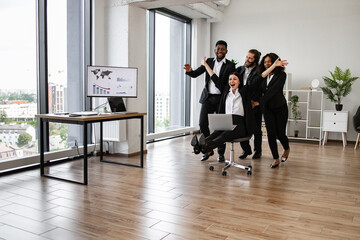 Business team in suits enjoying playful moment in office. Group of colleagues with woman on rolling chair showing teamwork and fun at work. Bright office space with large windows.